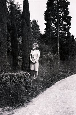 A woman in a knee-length dress and jacket stands beside a gravel path and tall, neatly-trimmed cypress trees. The setting app...