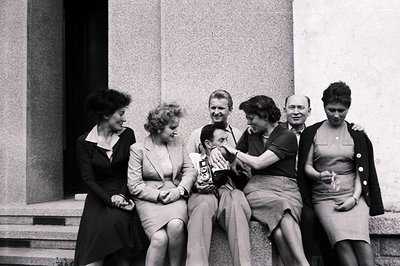 Six individuals are seated on wide stone steps; a man center holds a vintage camera, playfully interacting with a woman who r...