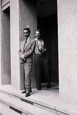 Two men in formal attire pose outdoors, framed by a modernist building's entrance. One wears a suit and tie, the other a shir...