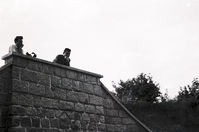 Two men in 1960s attire stand atop a stone retaining wall, seemingly engaged in conversation or observation. The wall leads u...