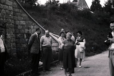 Group portrait; men in suits and casual attire converse with women in 1950s dresses near a stone retaining wall and hillside....
