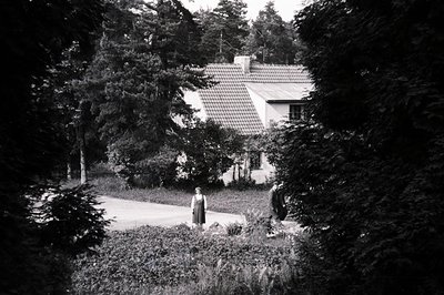 Framed by foliage, a charming, tile-roofed house sits within a lush garden. Two figures in long dresses stand on a driveway l...