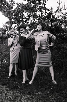 Three women in stylish 1960s suits pose amidst foliage. The group exudes confidence with dramatic stances and visible heels. ...