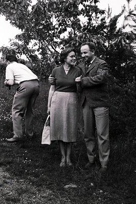 A couple stands in a garden, the man with his arm around the woman; a third figure is partially visible behind them. The scen...