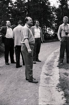 Group of men in suits & casual wear converse on a rural road. The man in the checkered shirt appears to be gesturing. Visible...