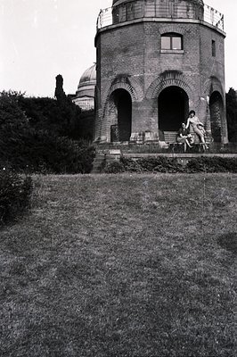 A cylindrical brick structure with arched openings sits atop a slight rise, two figures perched on the base. Dense foliage fo...