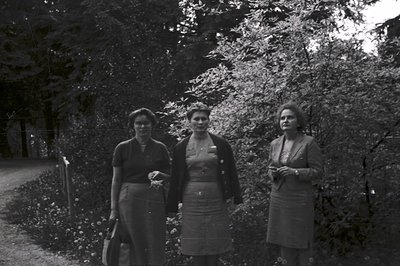 Three women stand in a garden, depicted in a black and white portrait. The composition suggests a candid moment; they are dre...