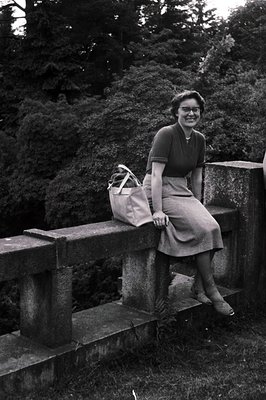 A smiling woman sits on a stone bridge railing, handbag at her side. She wears a fitted dress or top and a midi-length skirt,...
