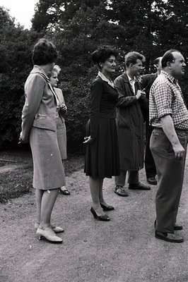 Group portrait of five people in a garden setting. Women wear 1960s-era tailored suits and dresses; men wear coats and plaid ...