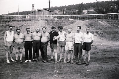 Group portrait of nine men, likely a volleyball team, in a sparse, outdoor setting. They wear athletic shorts and tank tops; ...