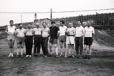 A group of eight men stand beside a volleyball net. Several are shirtless, all wear athletic shorts. A small building with a ...
