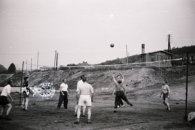 A group plays volleyball on a dirt court. Men in shorts and light-colored shirts compete. A makeshift net divides the space; ...