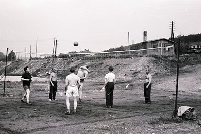 Five men play volleyball on a makeshift court with a wire net. Visible are simple athletic shorts and tops; the background fe...