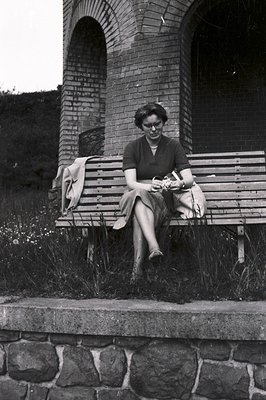 A woman in a dark skirt suit and glasses sits on a wooden bench beneath an arched brick structure. She holds a document or pa...