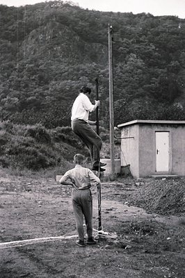 Two young men engage in a climbing game around a utility pole and small concrete building. One is ascending, the other observ...