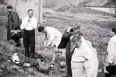 Group of men in suits and lab coats examine a body covered with a cloth on grassy terrain. Appears to be a forensic investiga...