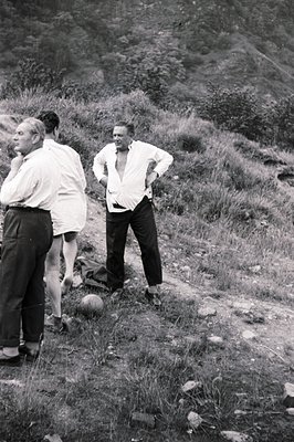B&W image: Two men in dress pants & open-necked shirts stand on a grassy slope with a small hill/mountain backdrop. One man i...