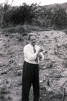 A full-length, black and white photograph depicts a man in a tailored suit and tie, gesturing outdoors on a sloped, grassy ar...