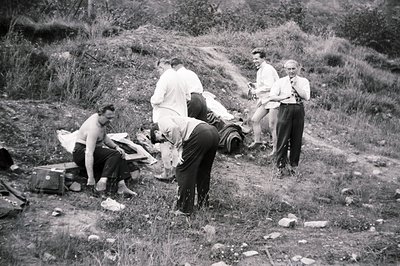 Group of men, dressed formally, appear to be exploring a rocky, overgrown hillside. Several suitcases and bags lie scattered ...