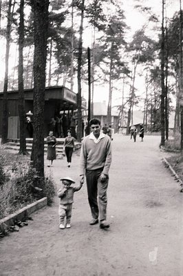 A father and young child walk along a paved path within a wooded area, likely a resort or recreational zone. The child wears ...