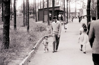 A young boy in a sailor suit walks alongside a man in a sweater, with another young girl following behind. A group of people ...