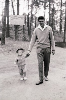 A man and young child walk hand-in-hand on a gravel path, framed by tall pine trees. The man wears a V-neck sweater, collared...