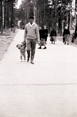A young man in a cable-knit sweater leads a toddler with a toy in a stroller along a paved path lined with birch trees. Sever...