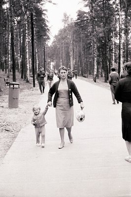 A woman walks along a paved path with a young child, both appearing lively. She wears a cardigan and knee-length skirt, holdi...