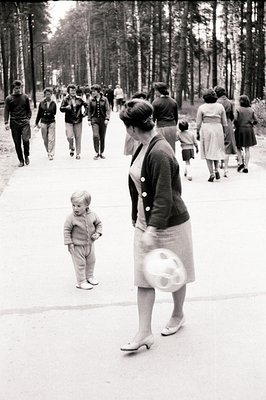 Black and white image shows a woman in a skirt suit & heels walking with a ball, a young boy looks back, amidst a group of pe...
