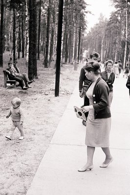 A candid street scene depicts a young boy taking tentative steps, a woman in a fitted skirt suit and heels approaching, and a...