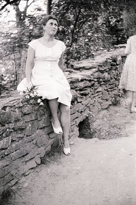 A young woman in a short, patterned dress and heels poses on a stone wall. A second figure, also in a dress, is blurred in th...