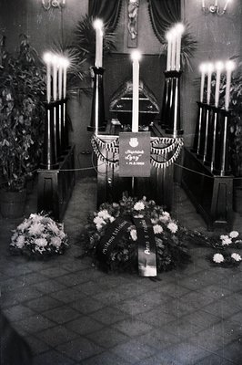 Solemn memorial scene within a church, featuring tall candelabras, floral arrangements, and a displayed plaque bearing text i...