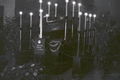 A formal, dimly-lit indoor scene with numerous tall, lit candles arranged around a draped table. A flag or banner with Cyrill...