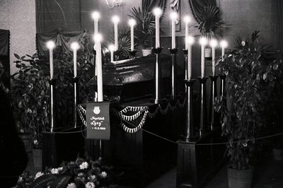 A tall, dark altar display features numerous lit candles, arranged within a multi-tiered structure and framed by potted plant...