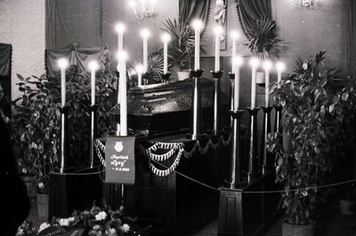 Elegantly draped casket displayed within a room, surrounded by tall, lit candles and potted greenery. A black banner with Cyr...