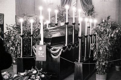Solemn memorial scene: A casket draped in fabric, encircled by tall, lit candles and potted plants. A banner bears Cyrillic s...