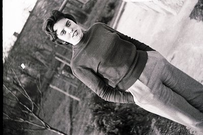 Young woman in a cable-knit sweater and high-waisted trousers stands outdoors, posing with a direct gaze. Architectural detai...