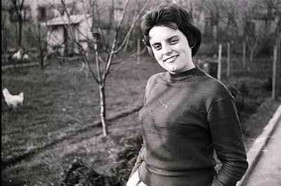 A smiling woman with a short, dark, bouffant hairstyle stands outdoors in a rural setting. She wears a dark, ribbed sweater a...