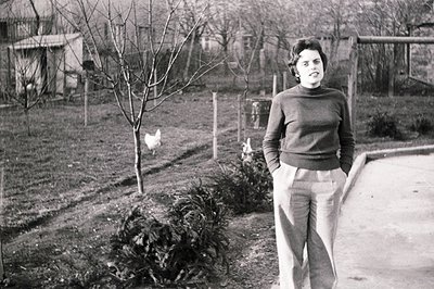 A young woman in a turtleneck sweater & trousers stands in a rural backyard, a chicken visible in the background. Simple fenc...