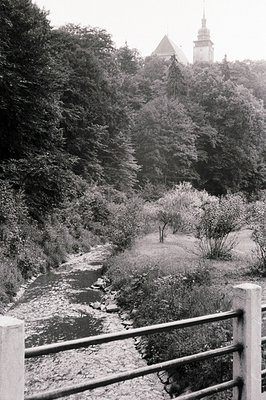 A river winds through a wooded valley, framed by a sturdy railing in the foreground. A stone structure with a spire rises abo...