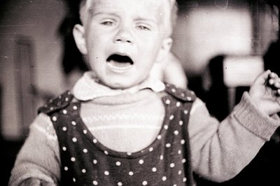 Close-up, black and white portrait of a young child crying with an open mouth. The child wears a patterned dress over a sweat...