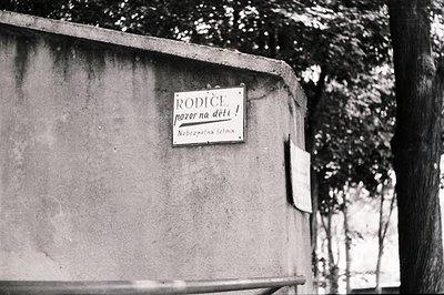 Close-up of a weathered sign attached to a concrete wall. The text, in Cyrillic script, reads “Rodīče, pozor na děti!” and “B...