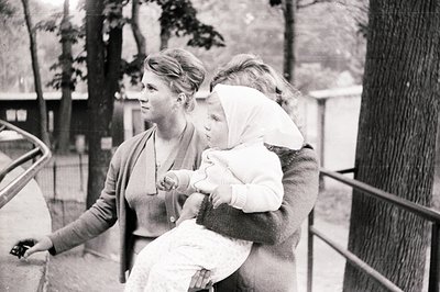 A woman in a cardigan holds a child bundled in a white bonnet, standing outdoors near a railing. The scene suggests a public ...