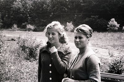 Two young women in cardigans stand near a park bench, gazing off-camera. Their hairstyles and clothing suggest a mid-20th cen...