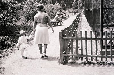 A woman and toddler walk along a paved path, hand-in-hand, next to a wooden picket fence. A seated figure rests on a bench vi...