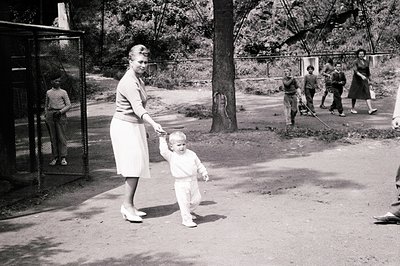 A mother and toddler walk hand-in-hand on a gravel path, likely in a park or zoo. The child, in overalls, takes unsteady step...