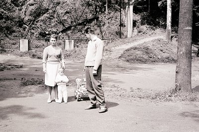 A young couple stands with a toddler outdoors, near a substantial bridge or viaduct structure. The woman wears a modest dress...