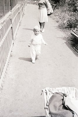 A young boy in a sweater and pants walks along a paved path. An adult in a skirt and carrying a bag walks away in the backgro...