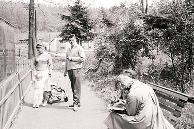 A young family strolls along a pathway, with a baby in a vintage pram. A woman sits on a bench, sketching in a notebook. Arch...