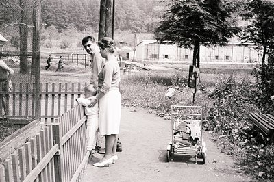 A man and woman, dressed in 1960s attire, observe a scene beyond a wooden fence, likely a zoo or park. The woman holds a youn...